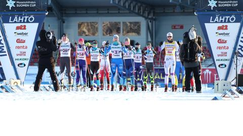 hiihtäjät rivissä lähtöpaikalla. Kaksi kameramiestä kuvaa hiihtäjiä.